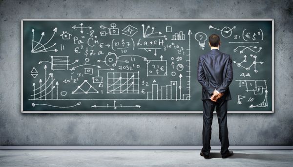 A professional in a dark suit stands in front of a blackboard covered with equations and graphs.